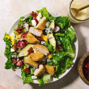 Cobb salad in a white bowl with chicken nuggets, dressing and other ingredients around it.