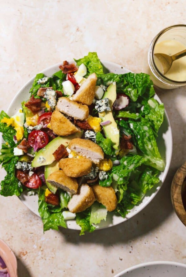 Cobb salad in a white bowl with chicken nuggets, dressing and other ingredients around it.