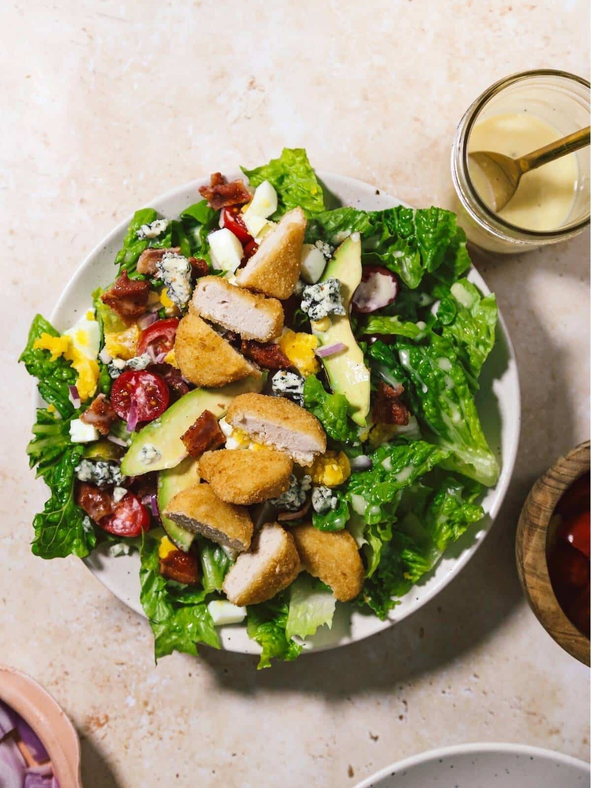 Cobb salad in a white bowl with chicken nuggets, dressing and other ingredients around it.