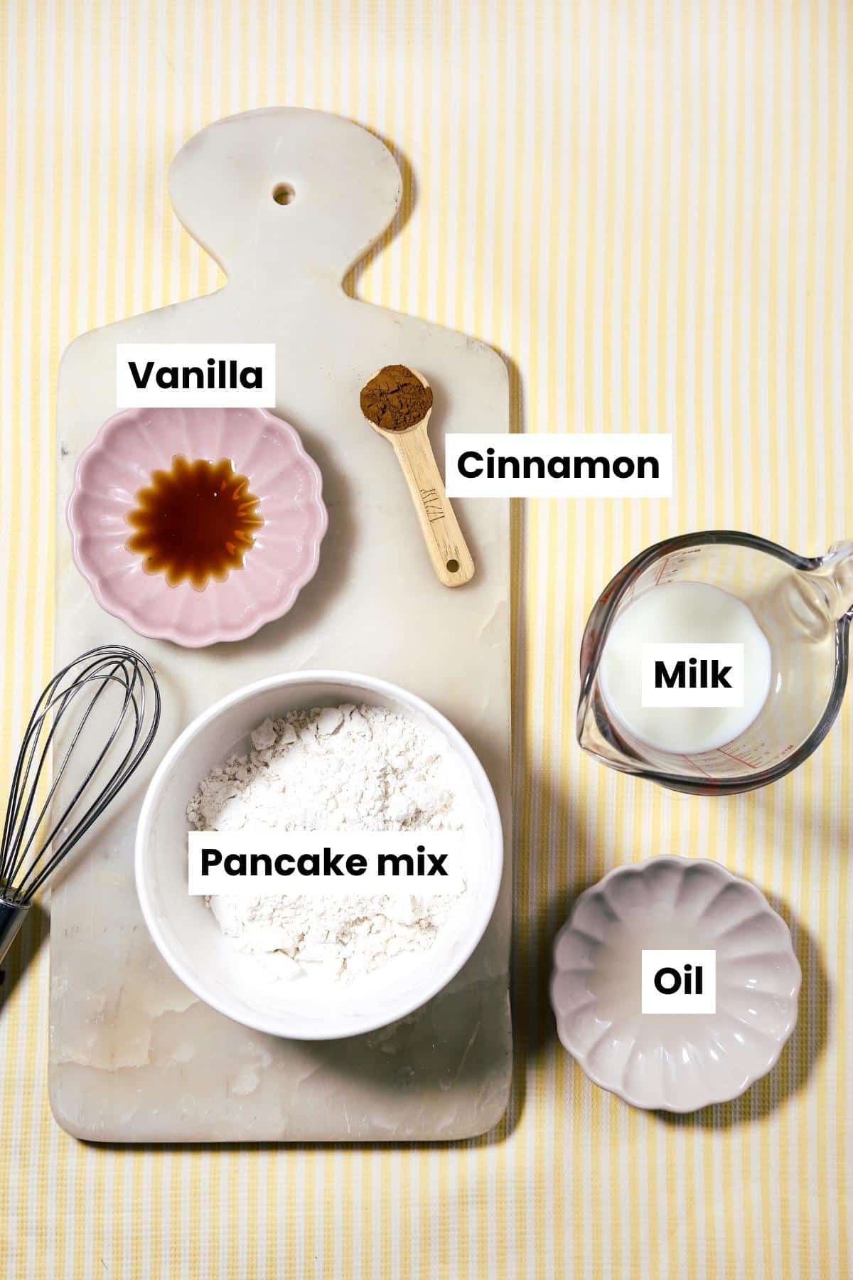 Ingredients and equipment laid out on stripped yellow background: vanilla, pancake mix, oil, cinnamon, milk, whisk and cutting board.