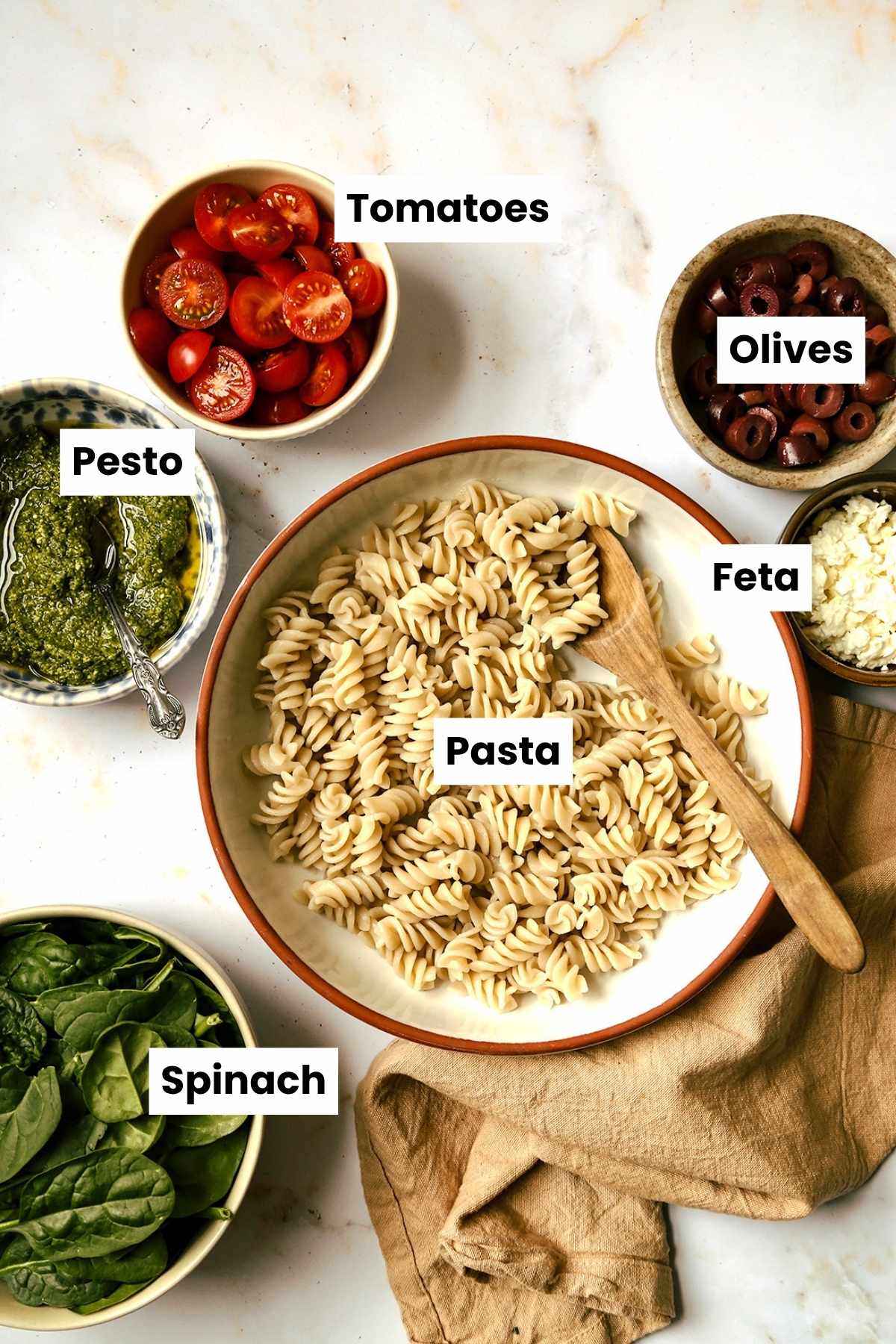 Overhead ingredients shot that includes pasta, feta, olives, spinach, pesto, and tomatoes.