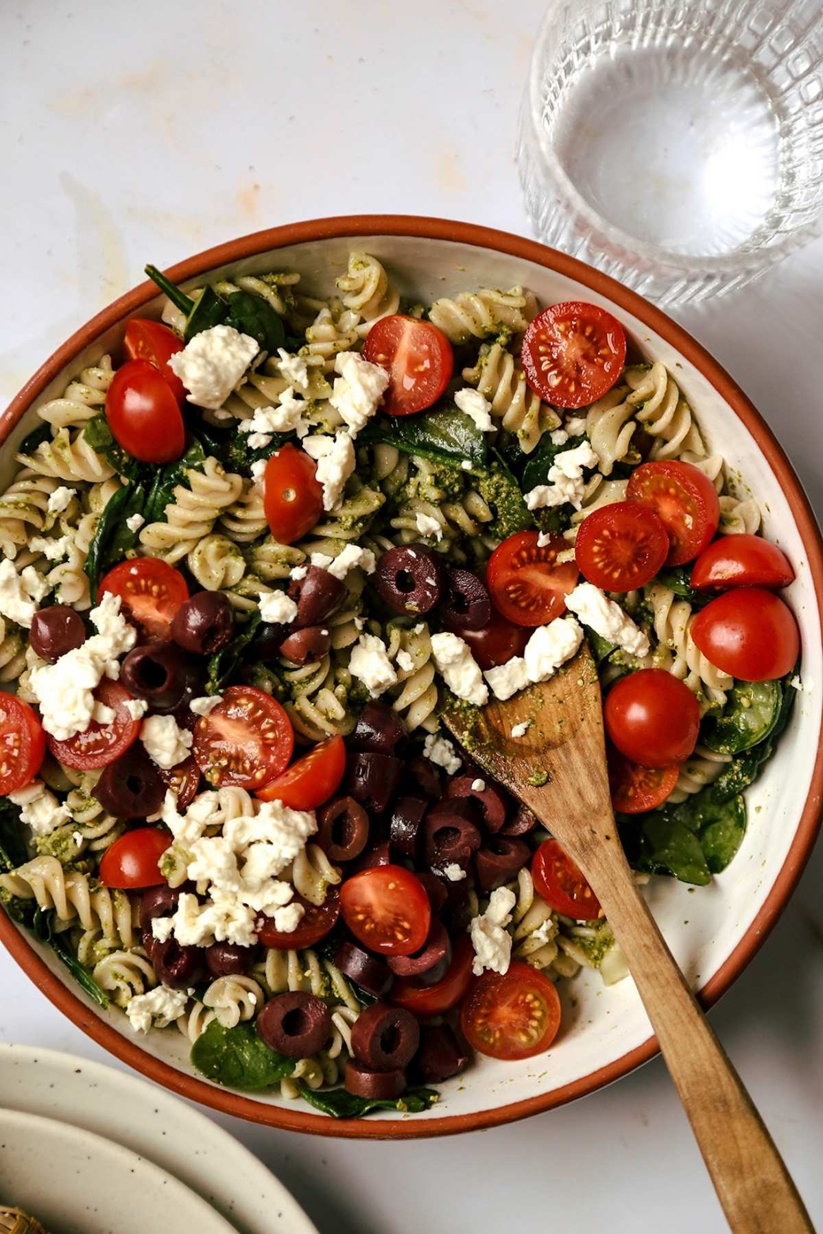 Pasta salad with pesto and feta in a bowl with a wooden spoon and a glass of water.
