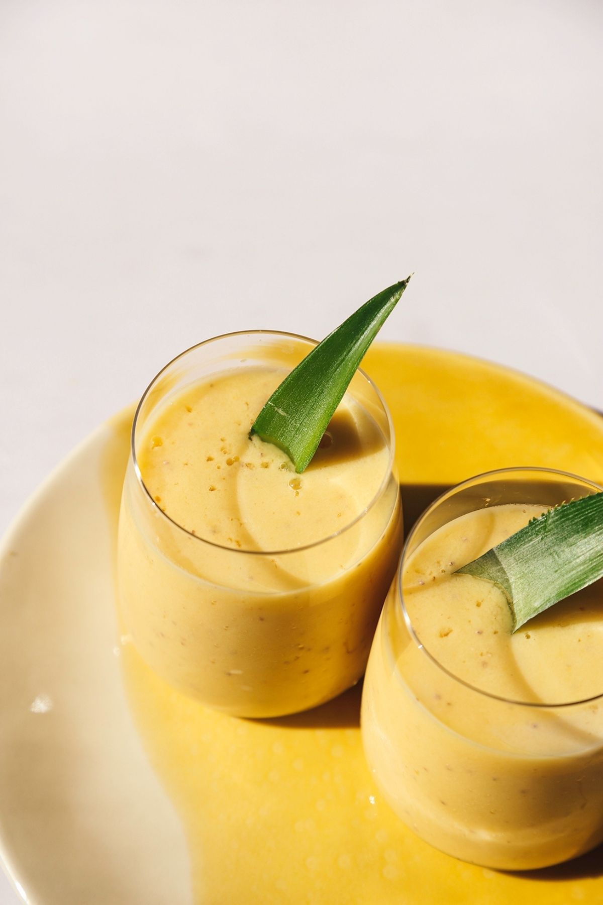 Image of two mango pineapple smoothies on a yellow plate with a pineapple leaf garnish.